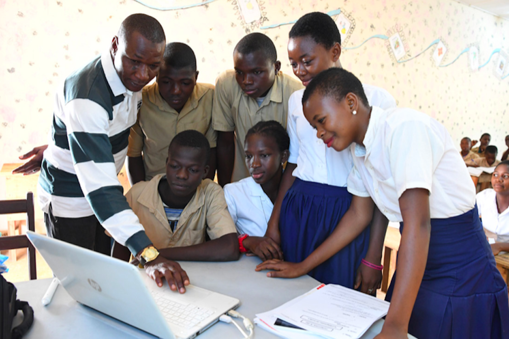 Gender-diverse group of Black children in school uniforms, having something demonstrated to them on a laptop in a classroom. The instructor is a Black man.