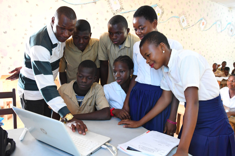 Gender-diverse group of Black children in school uniforms, having something demonstrated to them on a laptop in a classroom. The instructor is a Black man. Gender-diverse group of Black children in school uniforms, having something demonstrated to them on a laptop in a classroom. The instructor is a Black man.