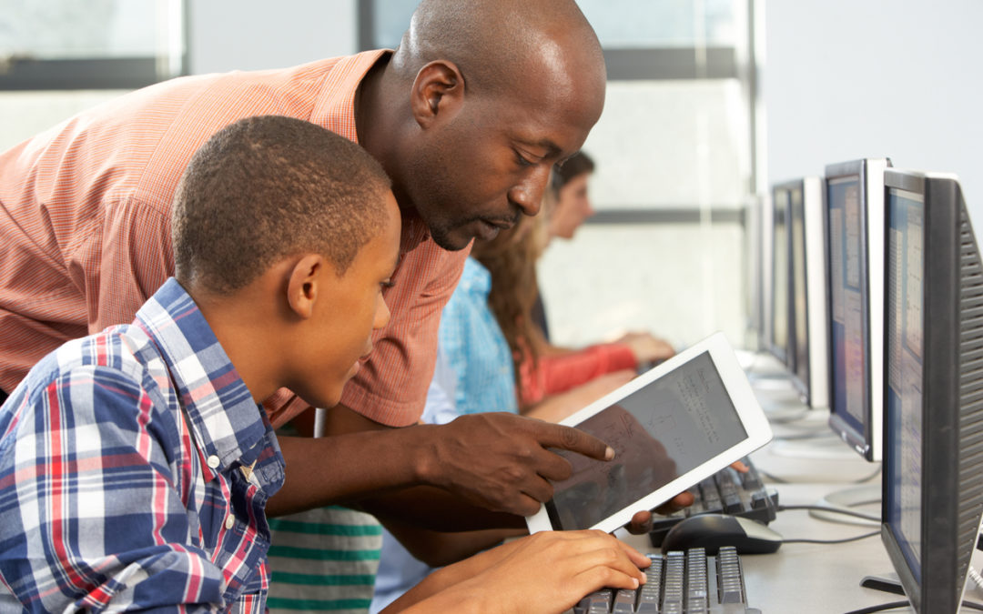 Teacher Helping Boy To Use Digital Tablet In Computer Class Instructor (who is a Black man) showing young Black boy something on a tablet, in a computer lab.